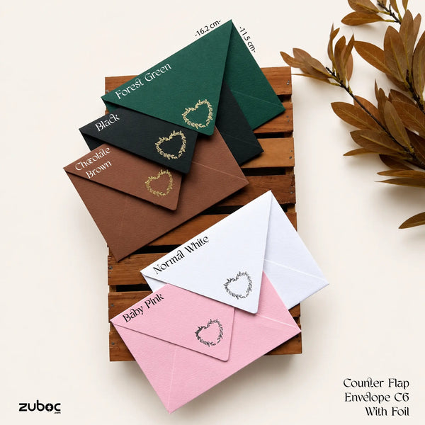 Six colored counter flap envelope with foil are arranged on a wooden slatted tray against a light beige background. The colors shown are Forest Green, Black, Chocolate Brown, Normal White, and Baby Pink.