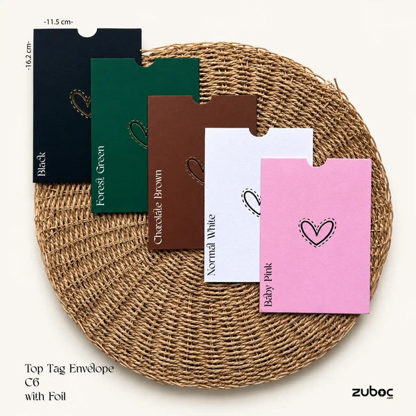 Five colored top tag envelopes with foil, displayed on a round woven mat. The colors shown are Black, Forest Green, Chocolate Brown, White, and Baby Pink, each with a different hand-drawn heart design.